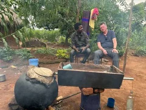 Two men sit beside a large drum still over a fire in a village yard, where kachasu is distilled.